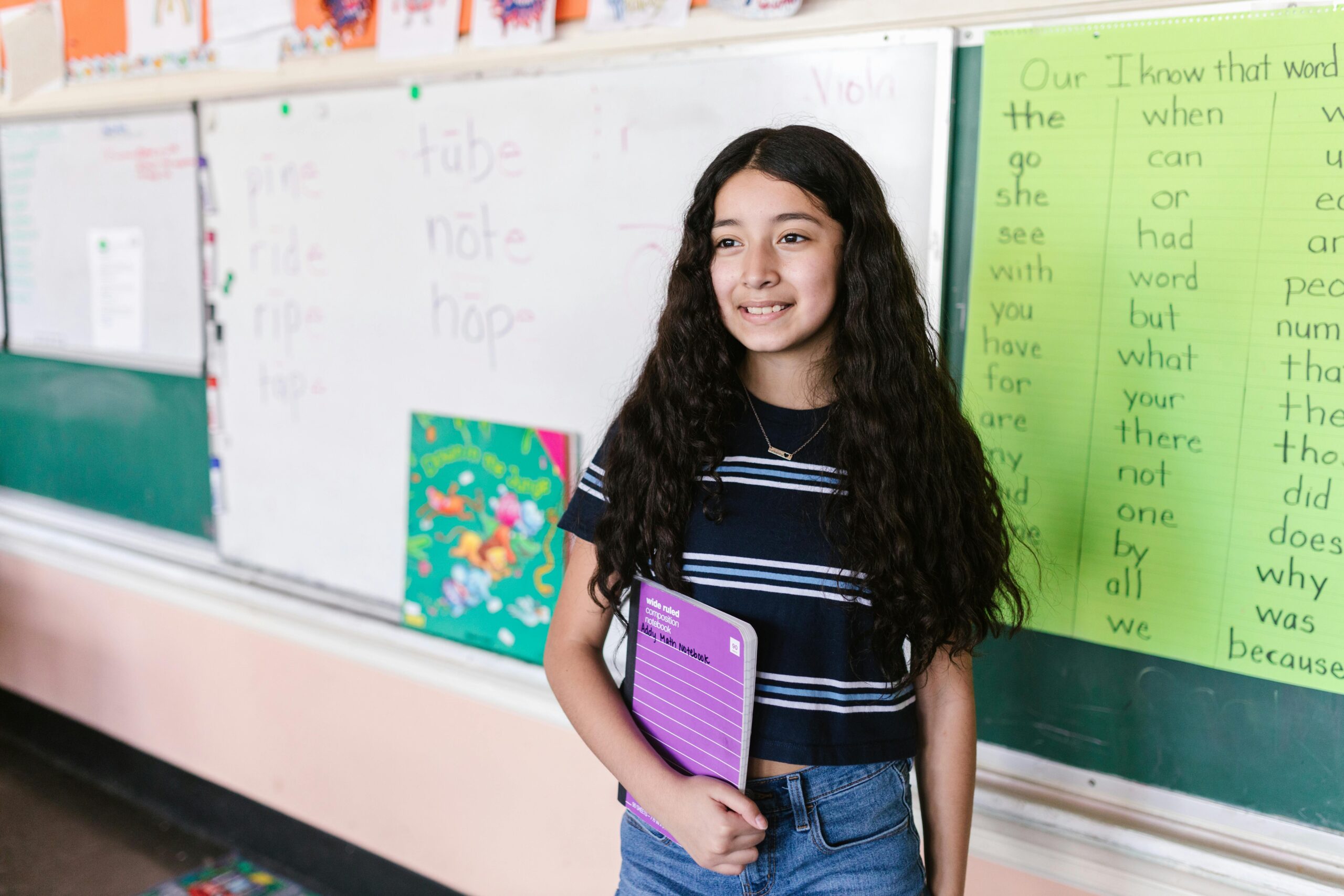 pexels-photo-8499551-8499551 Young girl in a classroom holding a notebook, smiling at the camera with a backdrop of educational materials.